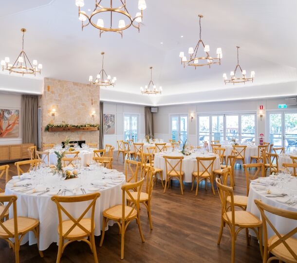 Bright event hall at The Homestead with round white-clothed tables, wooden chairs, chandeliers and abundant natural light