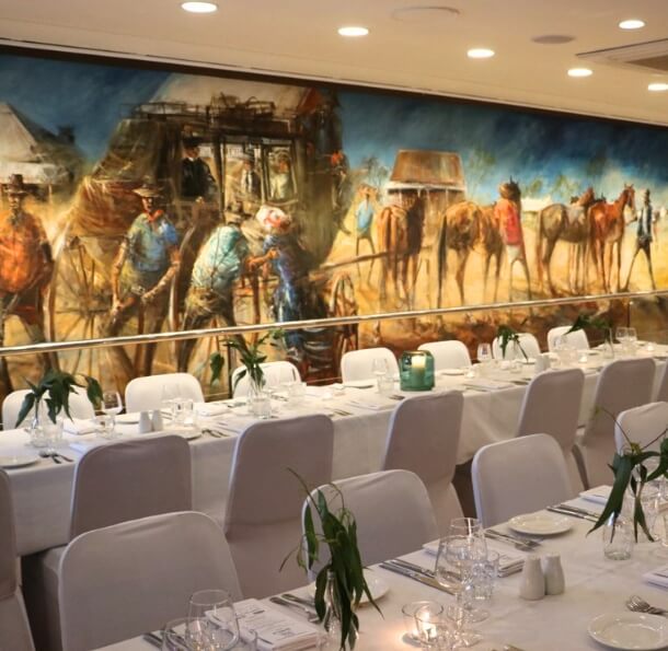 Banquet-style white tables set with candles and eucalyptus centerpieces in the Sawrey Room beneath a large cowboy mural