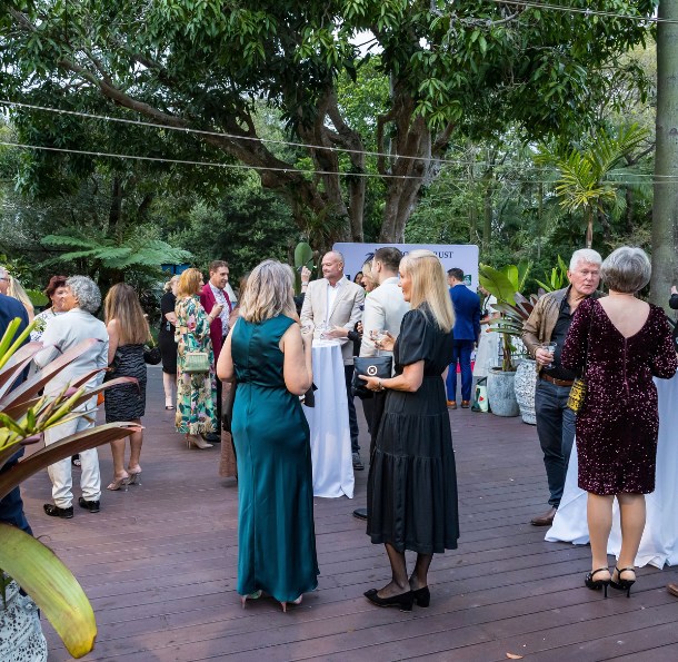Guests in elegant dresses and suits mingling at an outdoor cocktail reception on The Village Deck under large trees