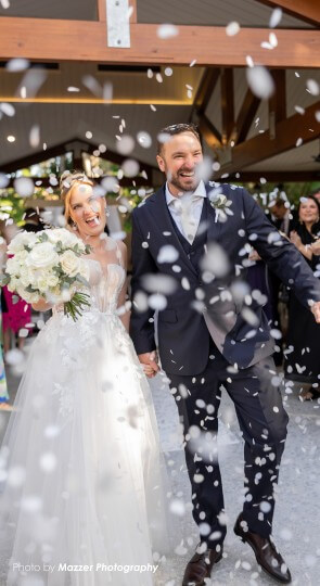 Bride in white gown and groom in dark suit walk down the aisle hand-in-hand, smiling as white confetti falls around them