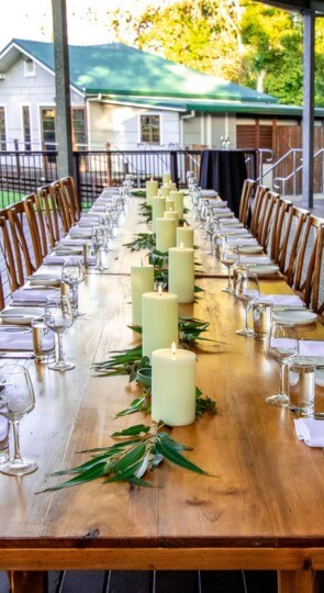 Long wooden table on a deck set for a meal with lit pillar candles, eucalyptus, wine glasses