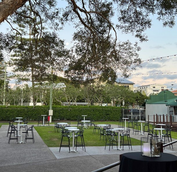 Outdoor deck with high cocktail tables and stools on concrete and grass, framed by hedges and a large tree with string lights
