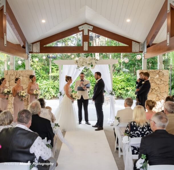 Bride and groom stand before an officiant under a white flowered arch in an open-air pavilion as seated guests look on