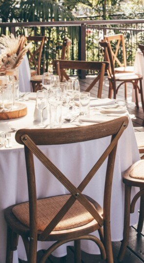 Outdoor veranda wedding setup: round tables with white linens, cross-back wooden chairs, glassware and simple centerpieces