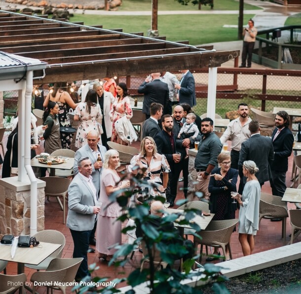 Guests in suits and long dresses mingling and holding drinks on an outdoor veranda under a wooden pergola at The Homestead