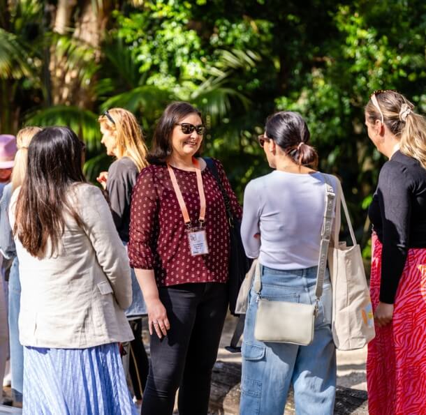 Group of women wearing lanyards chatting outdoors under trees at The Pavilion on a sunny day