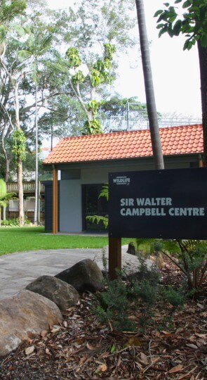 Tiled-roof building nestled among tall trees, paved path and rocks in front; sign reading Sir Walter Campbell Centre