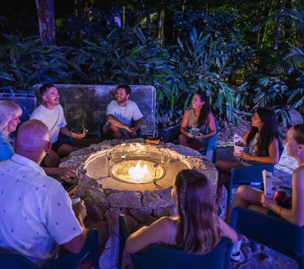 Group of people laughing and chatting while seated around an outdoor fire pit at night, lush plants in the background