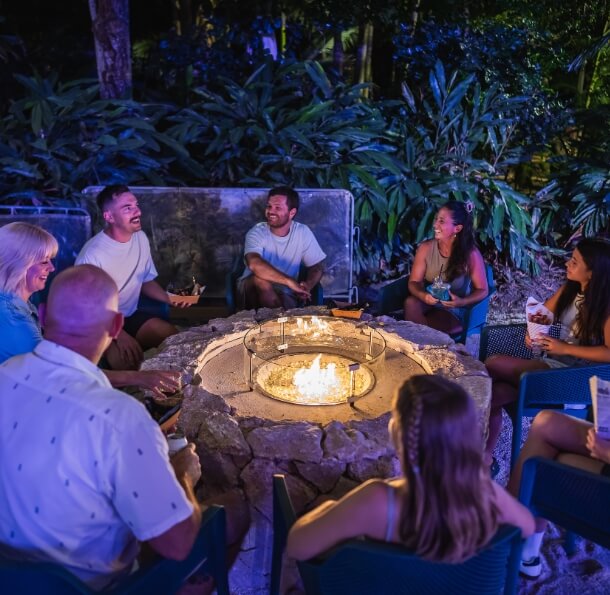 Group of people seated around a lit circular fire pit at night in a tropical garden, chatting and laughing