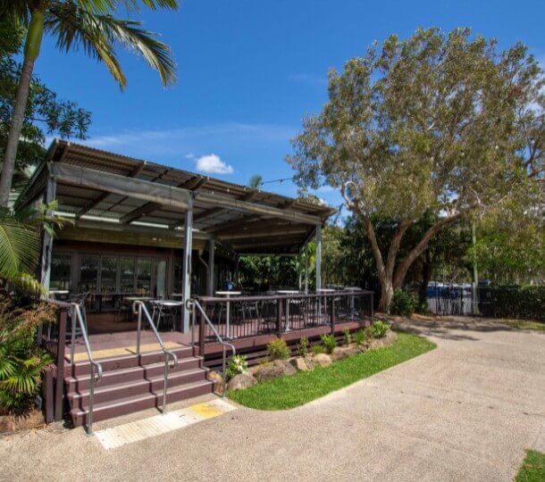 The Village Deck front with shaded outdoor veranda and seating, large trees, blue sky and concrete path with grass.