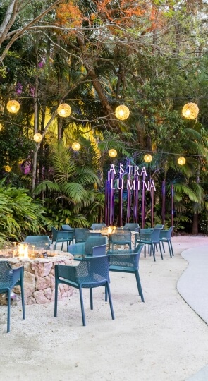 Turquoise chairs around lit fire pits under hanging globe lights, tropical trees, and illuminated sign reading Astra Lumina