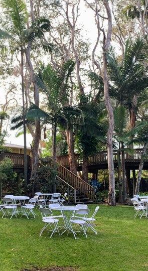 Outdoor lawn at Sir Walter Campbell Theatre with white folding tables and chairs beneath tall trees and a wooden deck.