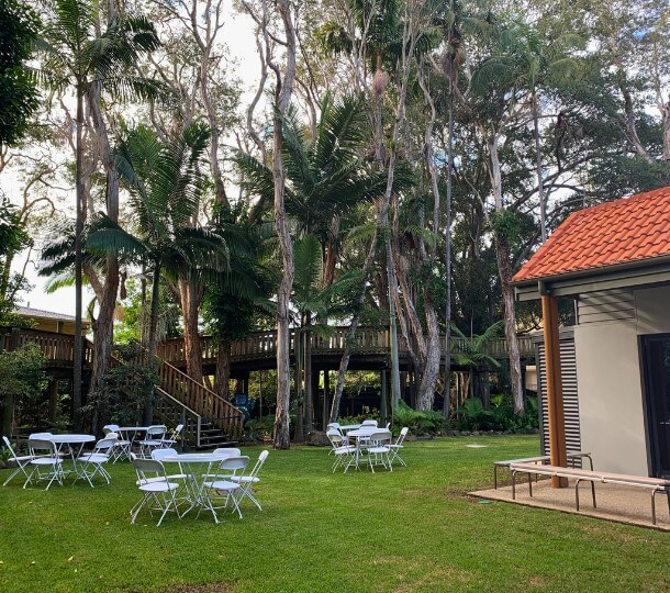 Outdoor lawn at Sir Walter Campbell Theatre with white folding tables and chairs under tall trees and a raised wooden deck.