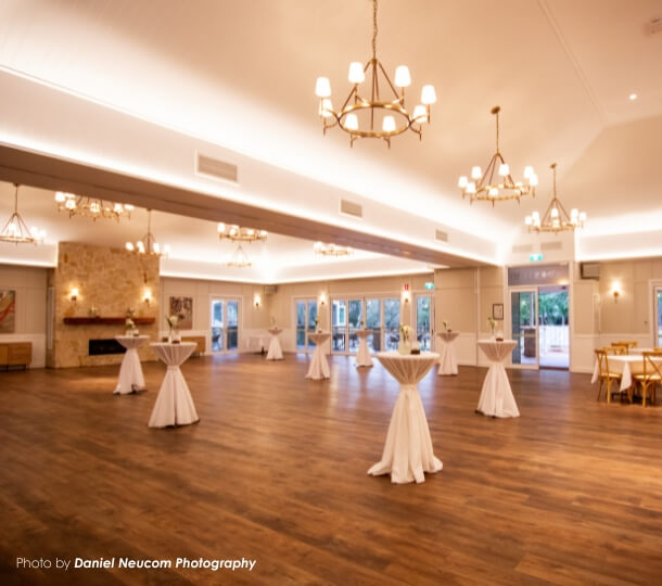 Spacious reception hall at The Homestead with wooden floor, warm chandeliers, wall lights and white-draped cocktail tables.