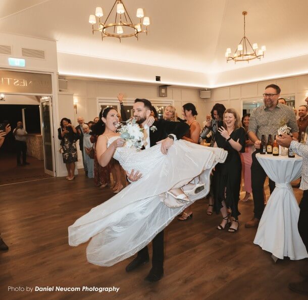 Groom carrying bride across a reception dance floor as cheering guests clap and smile, bride holding her bouquet.