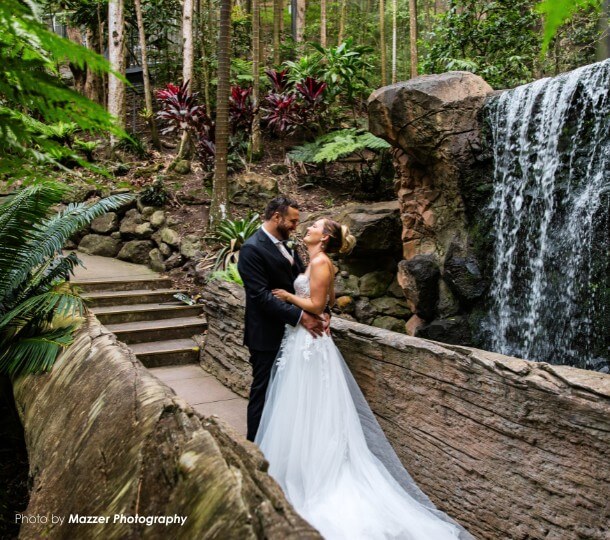 Bride in white gown and groom in suit embrace and smile on a natural rock bridge with waterfall in a tropical rainforest