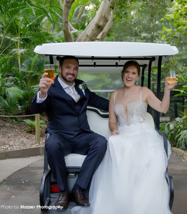 Newlywed couple in wedding dress and suit sitting in a golf buggy, smiling and raising drinks in a celebratory toast