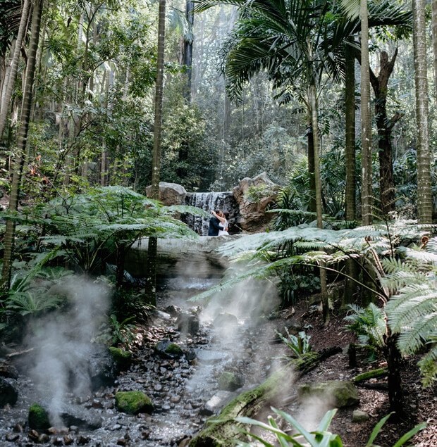 Couple embracing near a waterfall in a lush forest setting.
