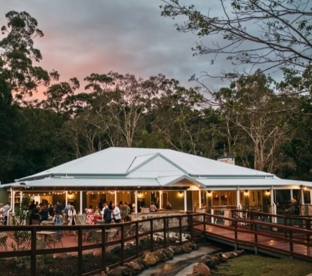 The Homestead — Queensland-style venue at sunset, people gathered on a large lit veranda surrounded by tall green trees