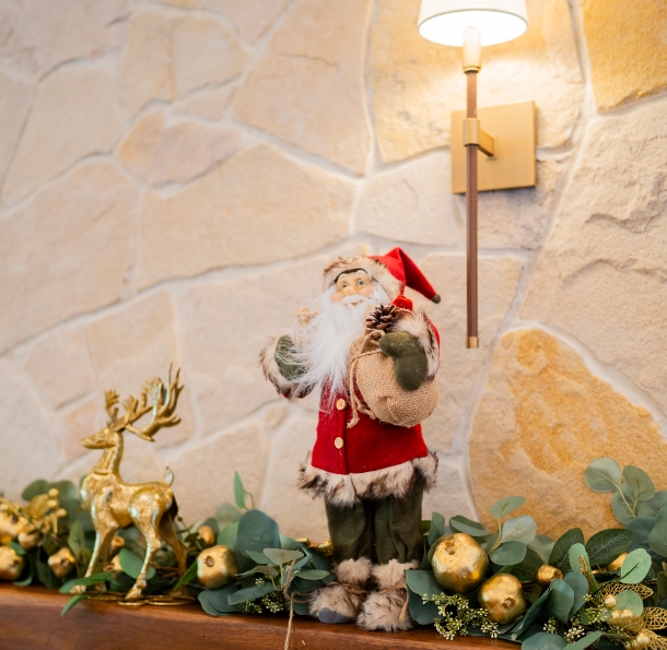 Christmas decorations on a wall shelf with a Santa Claus, golden reindeer, golden baubles and eucalyptus leaf strings. The wall shelf is attached to a modern rock wall fitted with a warm light lamp