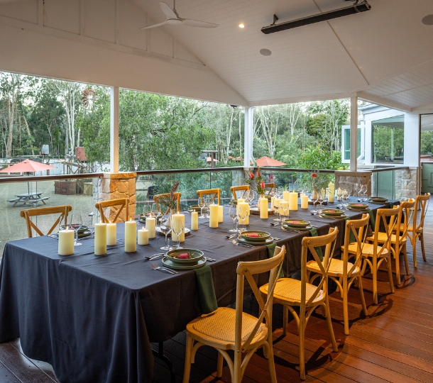 A long table on an outdoor undercover verandah overlooking natural bushland. The table is set with a black table cloth, candles, native Australian flower centre pieces and green dining ware decorated with red native flowers