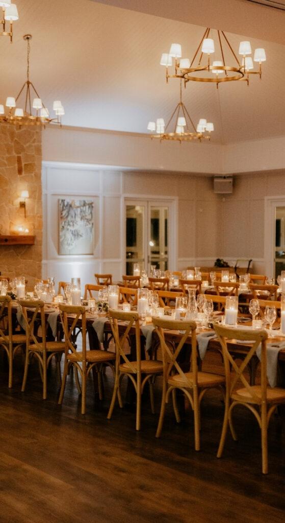 Elegantly set long tables with a white table runner, candles and glassware inside a rustic modern venue with chandeliers, large windows and warm lighting