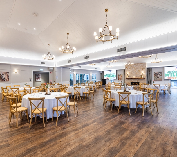Elegant modern rustic indoor venue with round tables, high ceilings and modern chandeliers. There are two large presentation screens on the back walls, nature-inspired paintings and a a gasfire