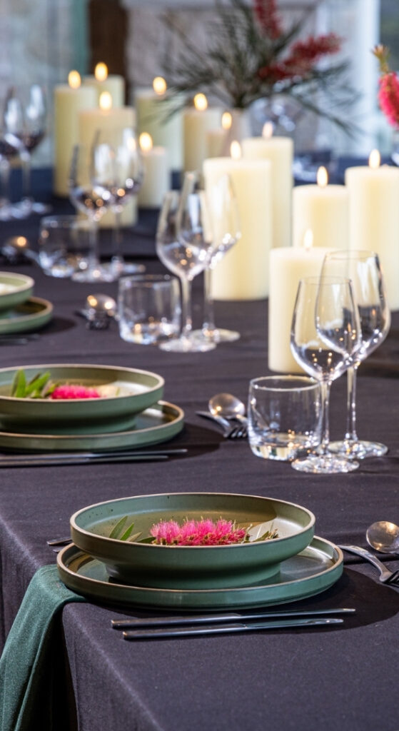 A closeup shot of a beautiful table set with lots of candles, glassware and green dining ware decorated with red native flowers.