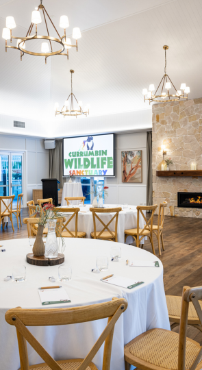 Elegant indoor tables set with a white table cloth, water glassware, centre piece and a pen and paper, inside a modern, well lit venue. The back wall features a large presentation screen and gas fire