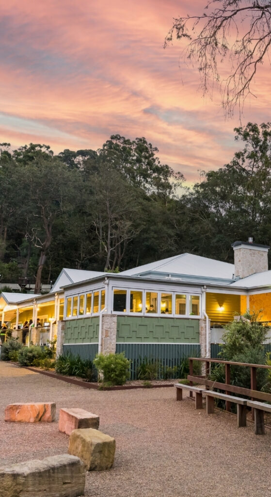 Sunset view of a green-and-white building surrounded by trees, with outdoor seating and a gravel pathway in front.