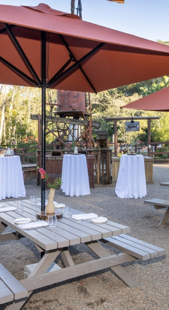 Rustic outback-style outdoor space with two large tables with burnt orange umbrellas and three tall cocktail tables set with a white tablecloth. The space has a farmyard barn and a large rustic wind mill, with large trees in the background