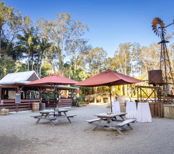 Rustic outback-style outdoor space with two large tables with burnt orange umbrellas and three tall cocktail tables set with a white tablecloth. The space has a farmyard barn and a large rustic wind mill, with large trees in the background