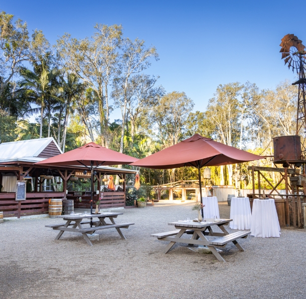 Rustic outback-style outdoor space with two large tables with burnt orange umbrellas and three tall cocktail tables set with a white tablecloth. The space has a farmyard barn and a large rustic wind mill, with large trees in the background