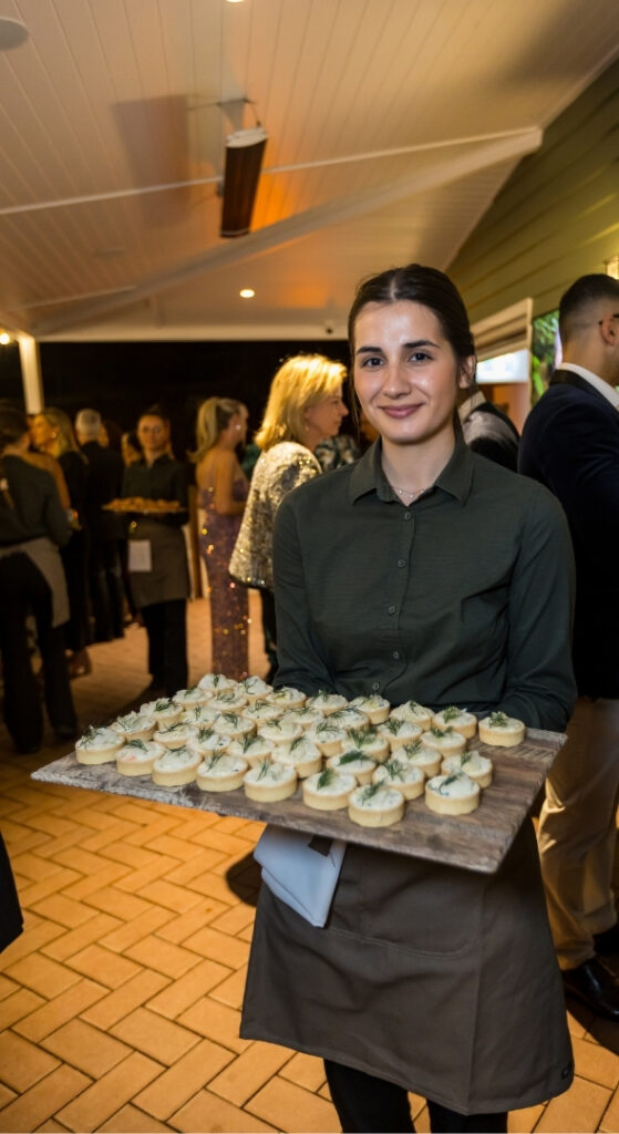 A server in a green shirt holds a wooden tray of canapés at a corporate event, with guests mingling in the background.