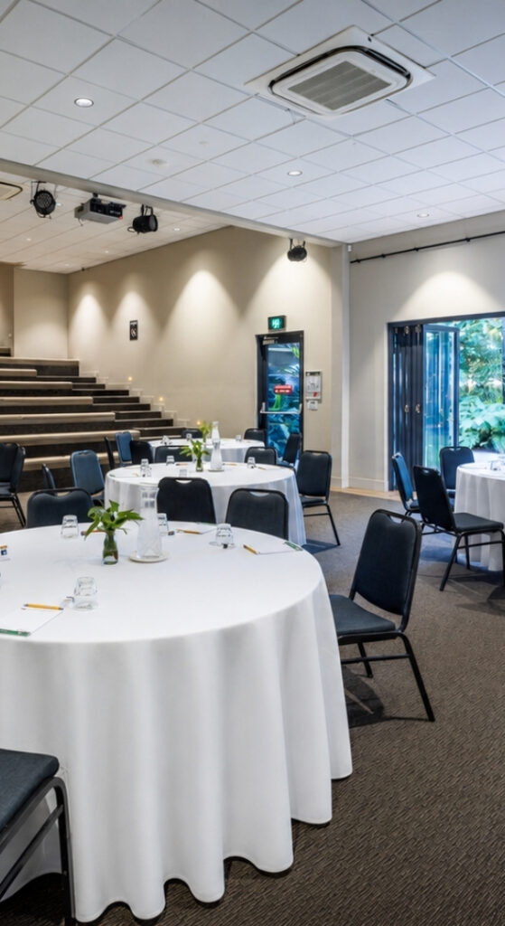 An indoor venue with round set tables and a large wall presentation screen with top to bottom large windows outlooking greenery. The back of the venue has tiered seating