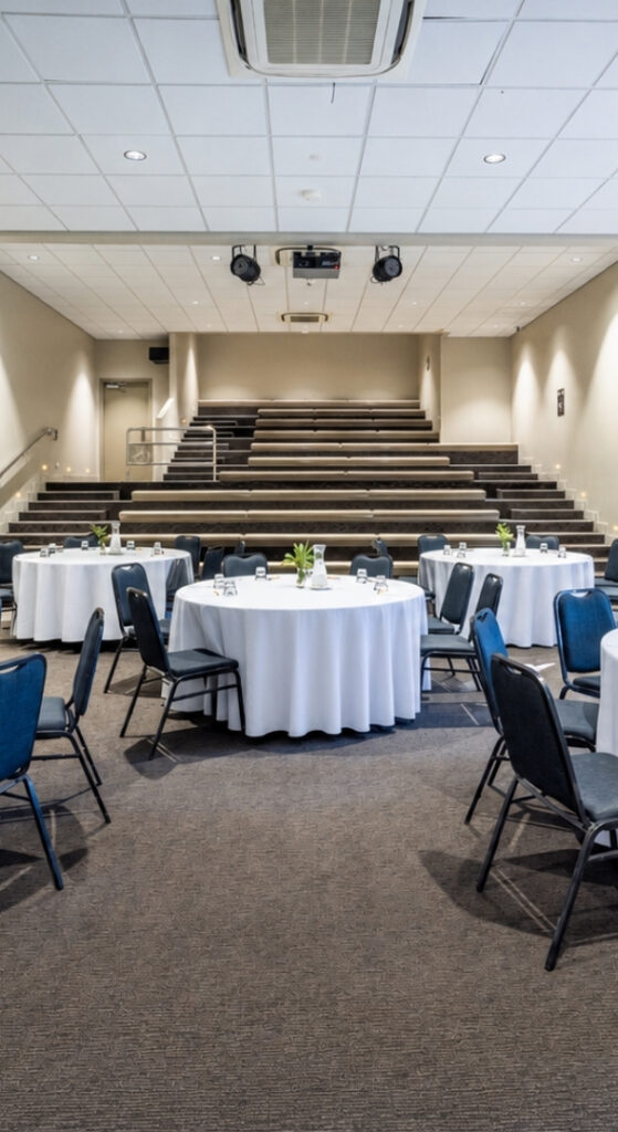 A large indoor venue with round set tables, wall spotlights, tiered seating and carpet with glimpses of large bottom to ceiling windows opening into an outdoor natural area