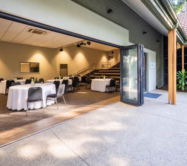 A large indoor venue with round set tables, wall spotlights, tiered seating and carpet with glimpses of large bottom to ceiling windows opening into an outdoor natural area