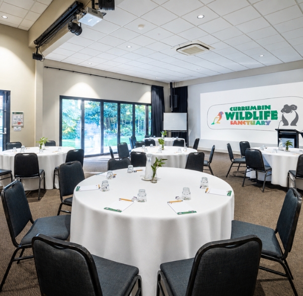 An indoor venue with round set tables and a large wall presentation screen with top to bottom large windows outlooking greenery