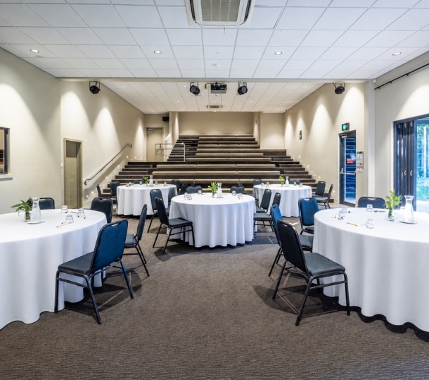 A large indoor venue with round set tables, wall spotlights, tiered seating and carpet with glimpses of large bottom to ceiling windows opening into an outdoor natural area