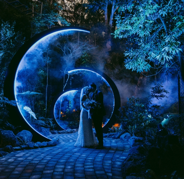 A wedding couple kissing underneath a circular moon installation with blue lights and fog effects at night