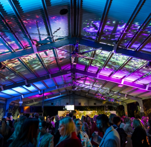 A large group of people networking at a open shed venue. The ceiling is illuminated with purple and blue lights.