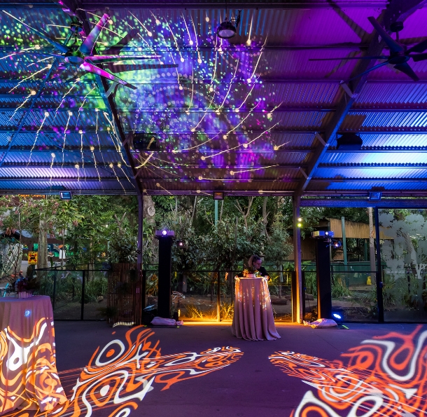 Colorful light projections cover the floor and ceiling of an indoor event space with tables and a DJ booth, surrounded by glass walls showing a garden outside.