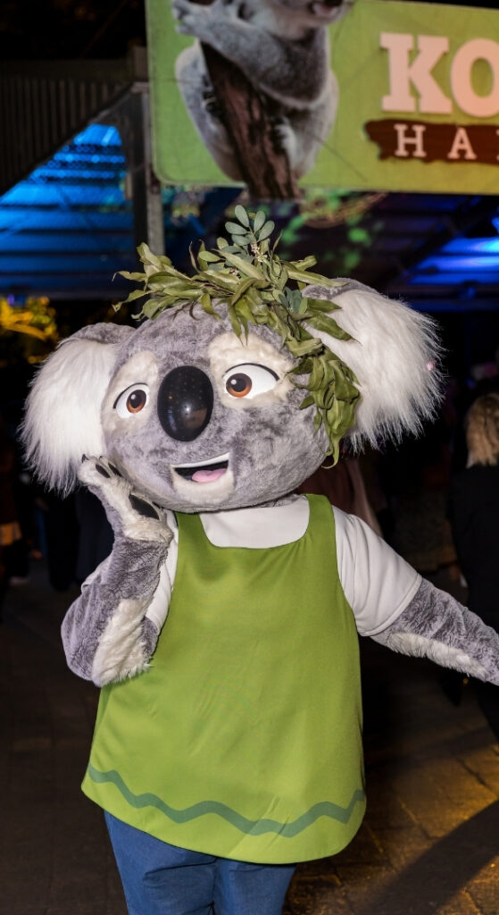 Koala mascot character wearing a light green vest and eucalyptus head crown, at night