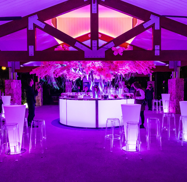 Cocktail bar setup under a pavilion with purple lighting, floral installation, and illuminated stools at a night event