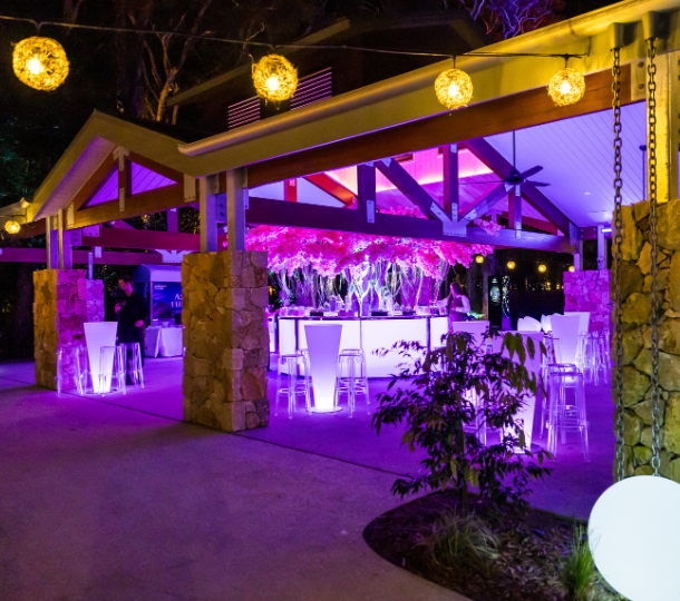 Pavilion cocktail event at night with purple lighting, floral installation, and illuminated stools
