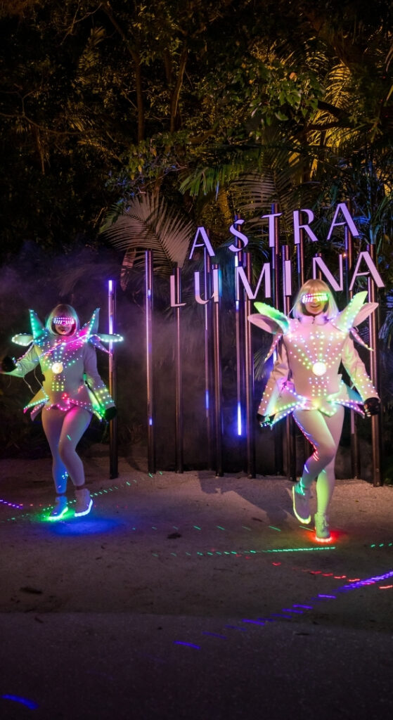Performers in illuminated LED costumes walking past the Astra Lumina sign at night