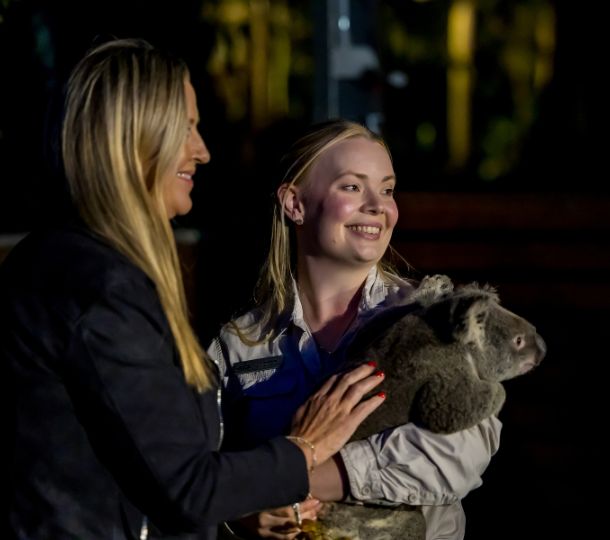 Two women stand together; one is holding a koala while the other is gently touching the animal. The setting appears to be outdoors at night.