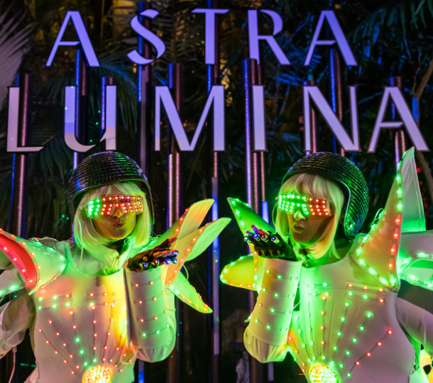 Performers in illuminated LED costumes posing in front of the Astra Lumina sign at night